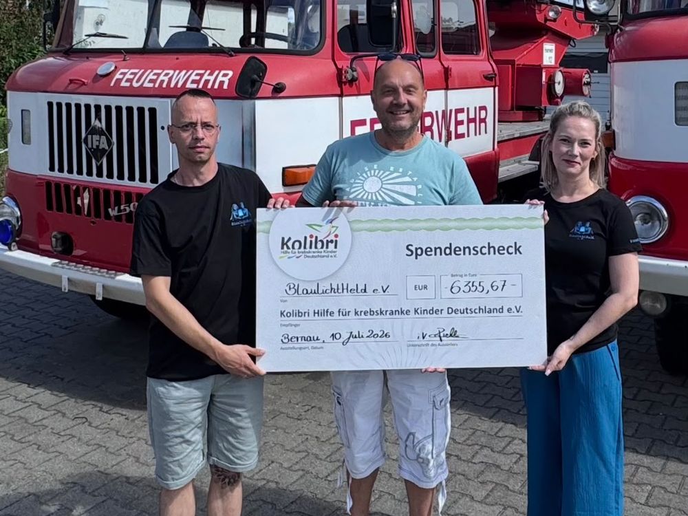 Drei Personen halten vor zwei historischen Feuerwehrfahrzeugen einen Spendenscheck über 6.355,67 Euro